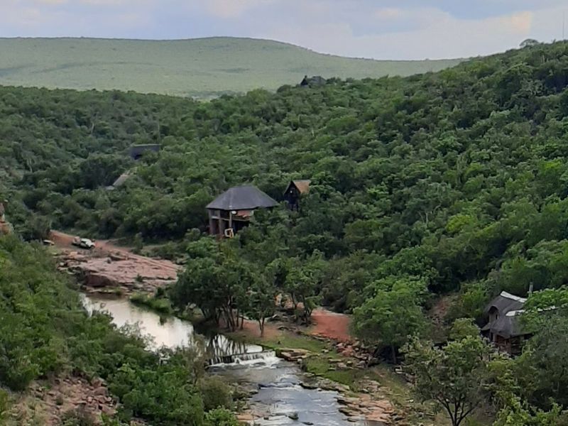 A scenic elevated view of a natural river flowing through a lush, wooded valley with secluded thatched-roof dwellings nestled in the landscape.