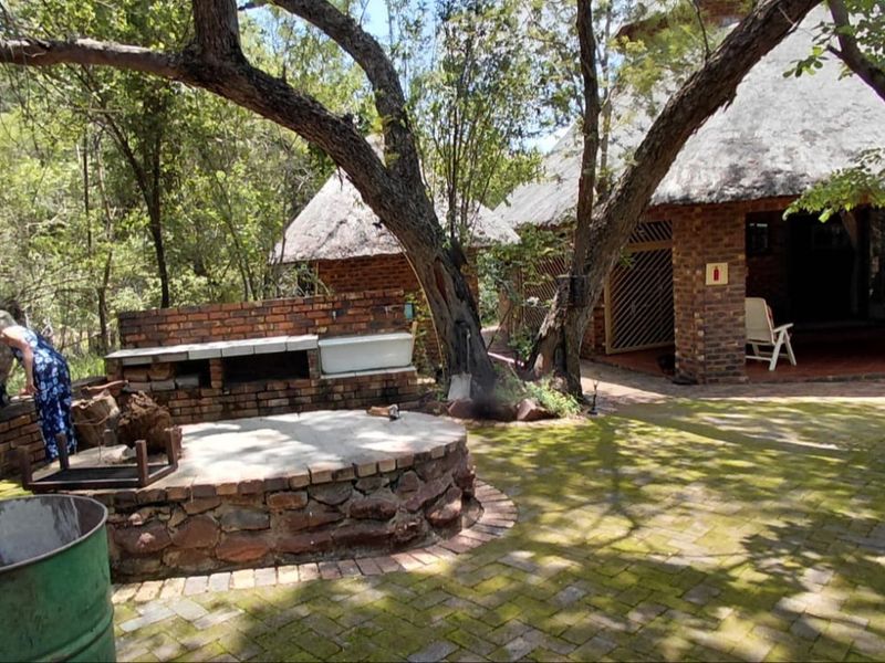 Outdoor patio area featuring a brick built-in braai, brick paving, and access to a thatched-roof structure in a natural, wooded setting.