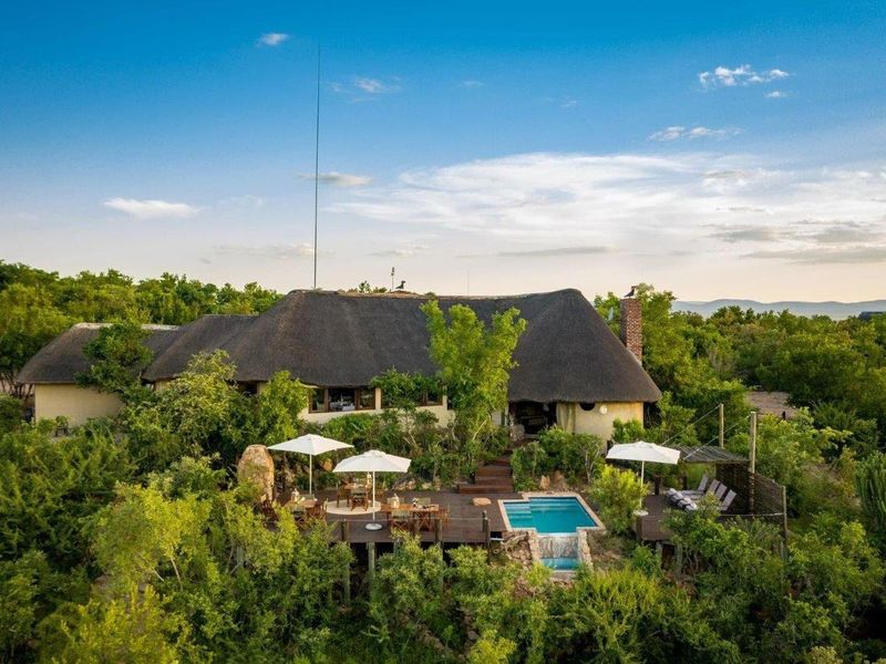 Aerial view of a farm-style residence featuring a traditional thatched roof, a raised timber deck, and a private swimming pool set in lush surroundings.