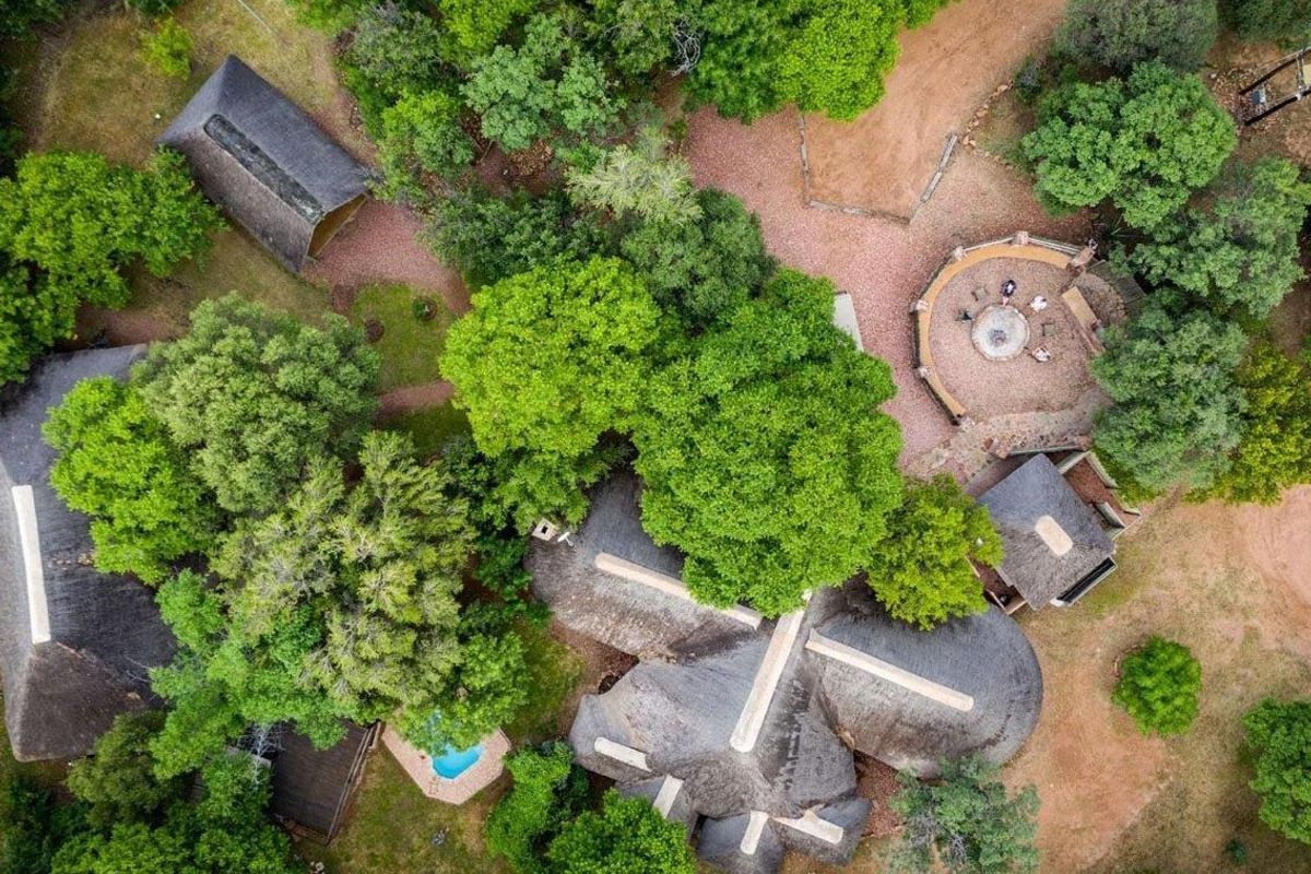 Aerial view of a farm property featuring multiple thatched-roof structures, a swimming pool, and a circular boma fire pit set within a wooded landscape.