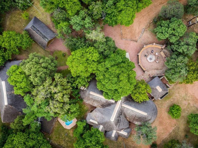 Aerial view of a farm property featuring multiple thatched-roof structures, a swimming pool, and a circular boma fire pit set within a wooded landscape.