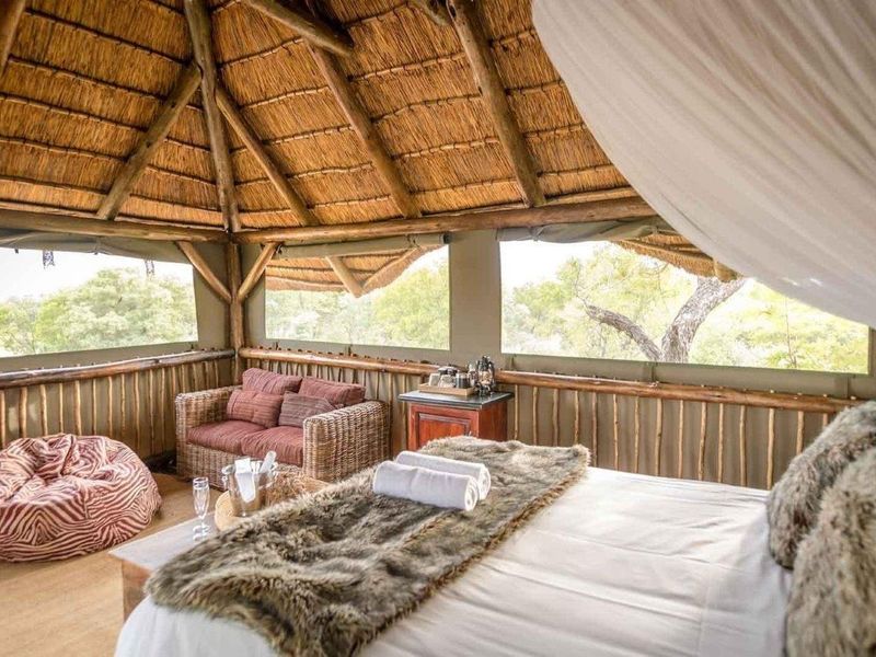 A rustic bedroom suite featuring a traditional thatched roof, exposed timber rafters, and panoramic canvas roll-up windows overlooking the surrounding landscape.