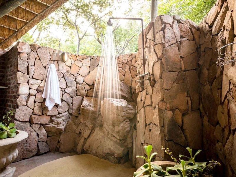 A unique outdoor shower area featuring natural stone wall cladding, a thatched roof overhang, and a rustic, integrated shower fixture.