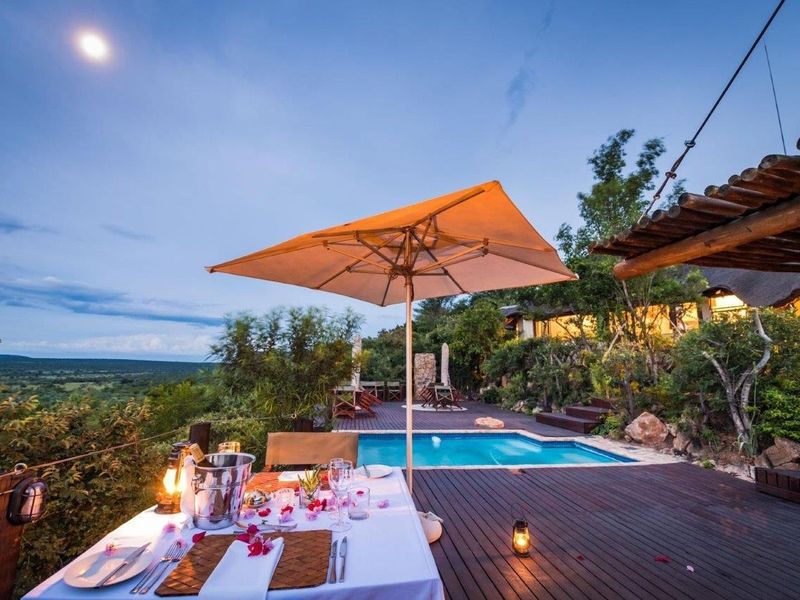 An inviting outdoor pool area featuring timber decking, a stone retaining wall, and a thatched roof structure overlooking a scenic landscape.