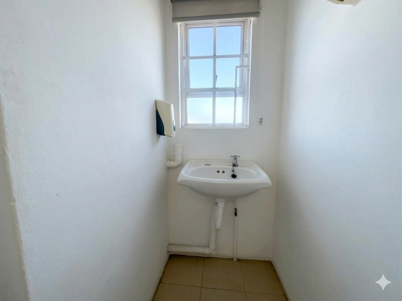 A small, functional utility space featuring a wall-mounted basin, tiled flooring, and a window with security bars.