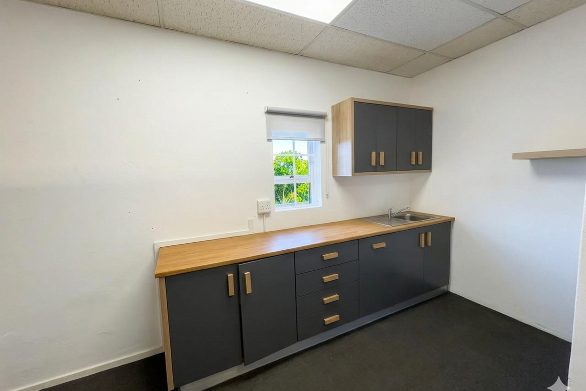 A clean, modern office kitchenette featuring dark grey cabinetry, wood-tone countertops, a stainless steel sink, and a drop ceiling with integrated lighting.