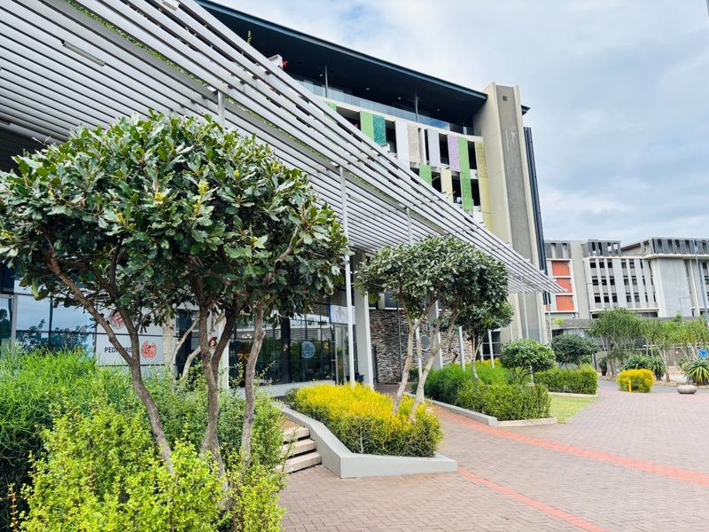 Modern commercial building exterior featuring a prominent louvred pergola, stone cladding accents, and well-maintained landscaped garden beds along a paved walkway.