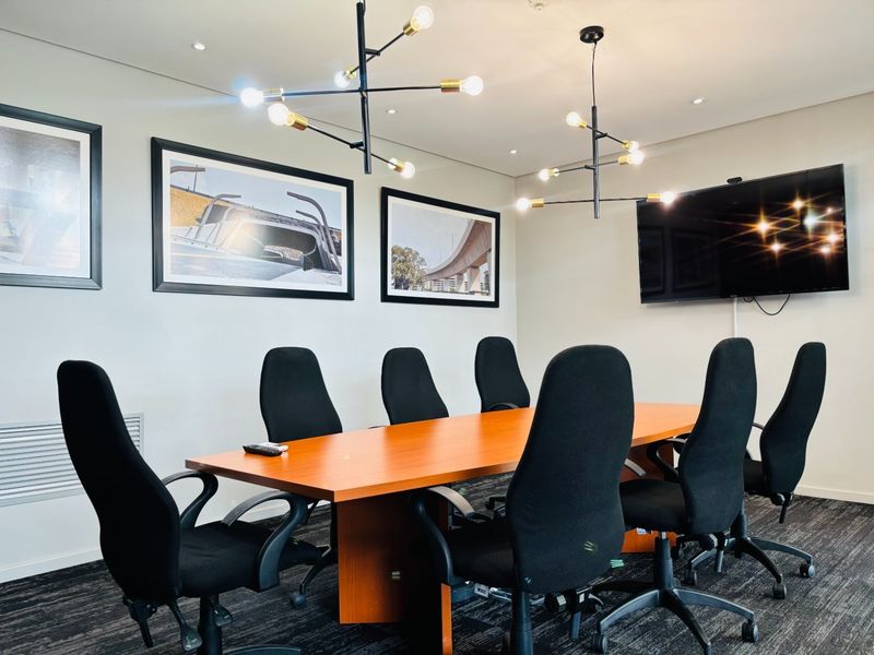 A professional commercial boardroom featuring modern pendant lighting, wall-mounted display screen, and carpeted flooring, suitable for corporate meetings.