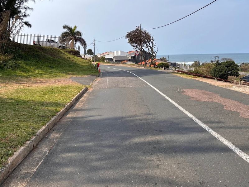 A street view showing the asphalt road, concrete curbing, and grass verge leading towards the coastal area.