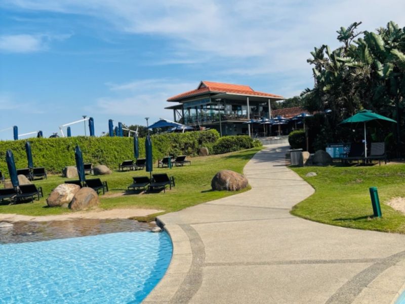 A well-maintained outdoor pool area featuring a paved walkway, landscaped gardens, and a clubhouse building in the background.