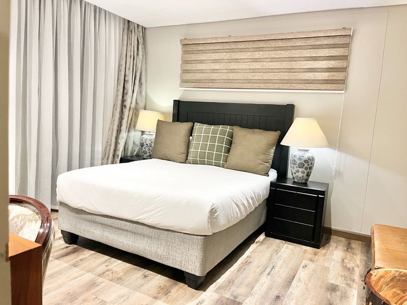 A well-maintained bedroom featuring modern laminate flooring, neutral wall finishes, and integrated window treatments including a roller blind and curtain tracks.