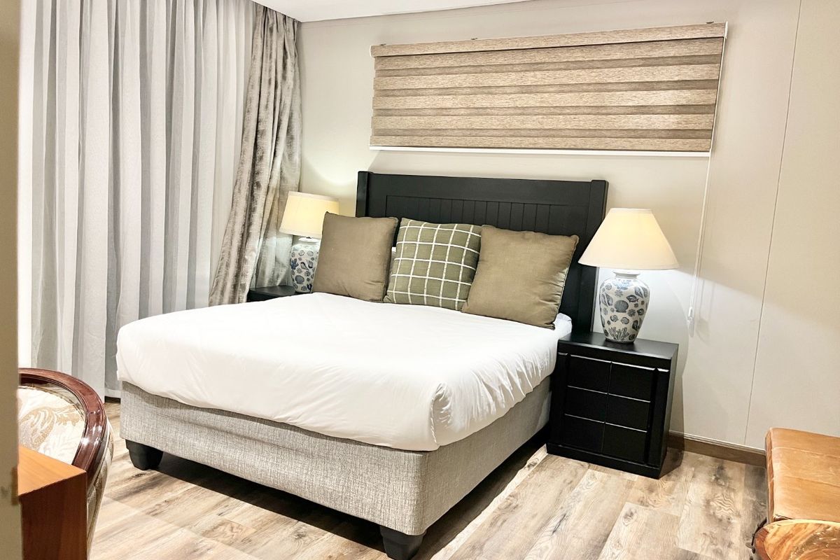 A well-maintained bedroom featuring modern laminate flooring, neutral wall finishes, and integrated window treatments including a roller blind and curtain tracks.