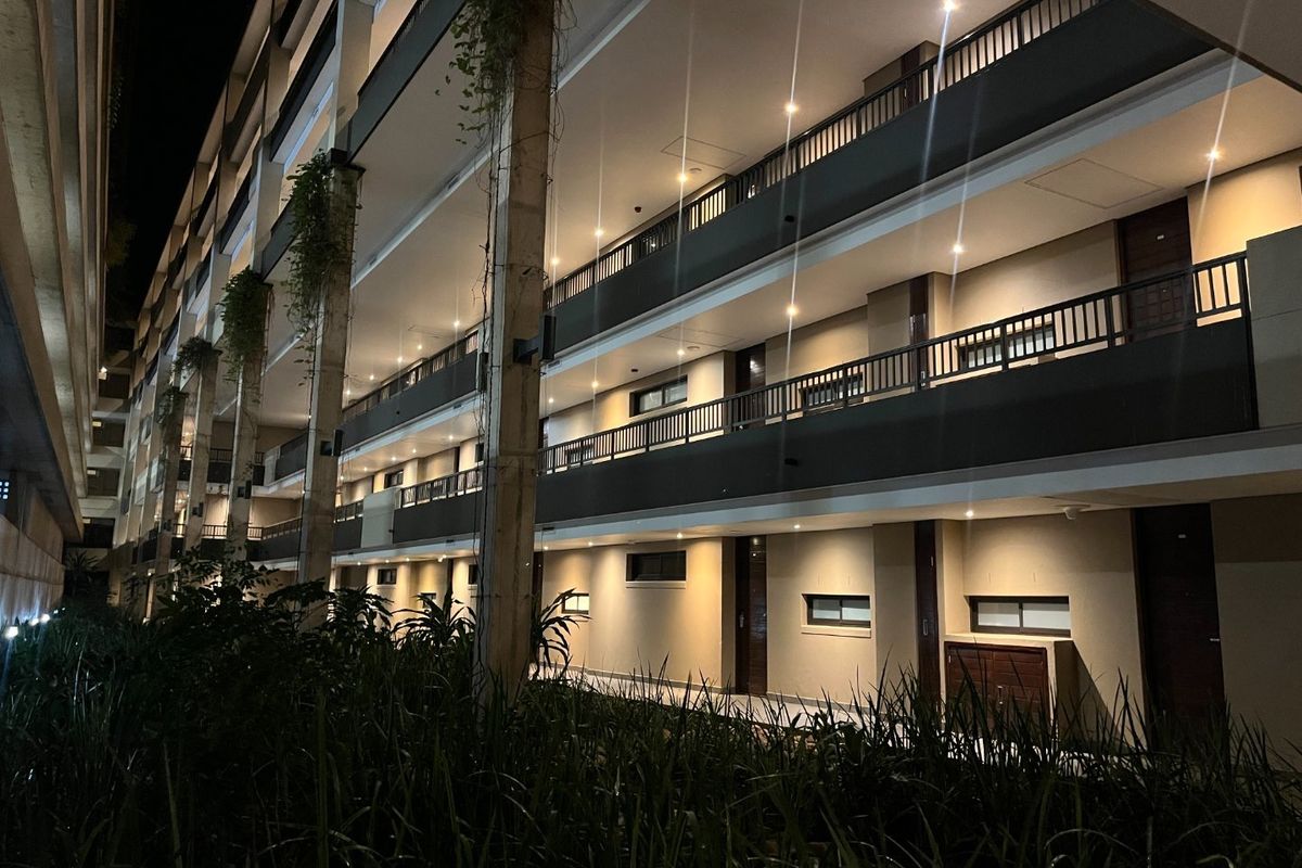 Night view of a multi-story apartment building exterior featuring illuminated balconies, metal railings, and landscaped ground-level garden beds.
