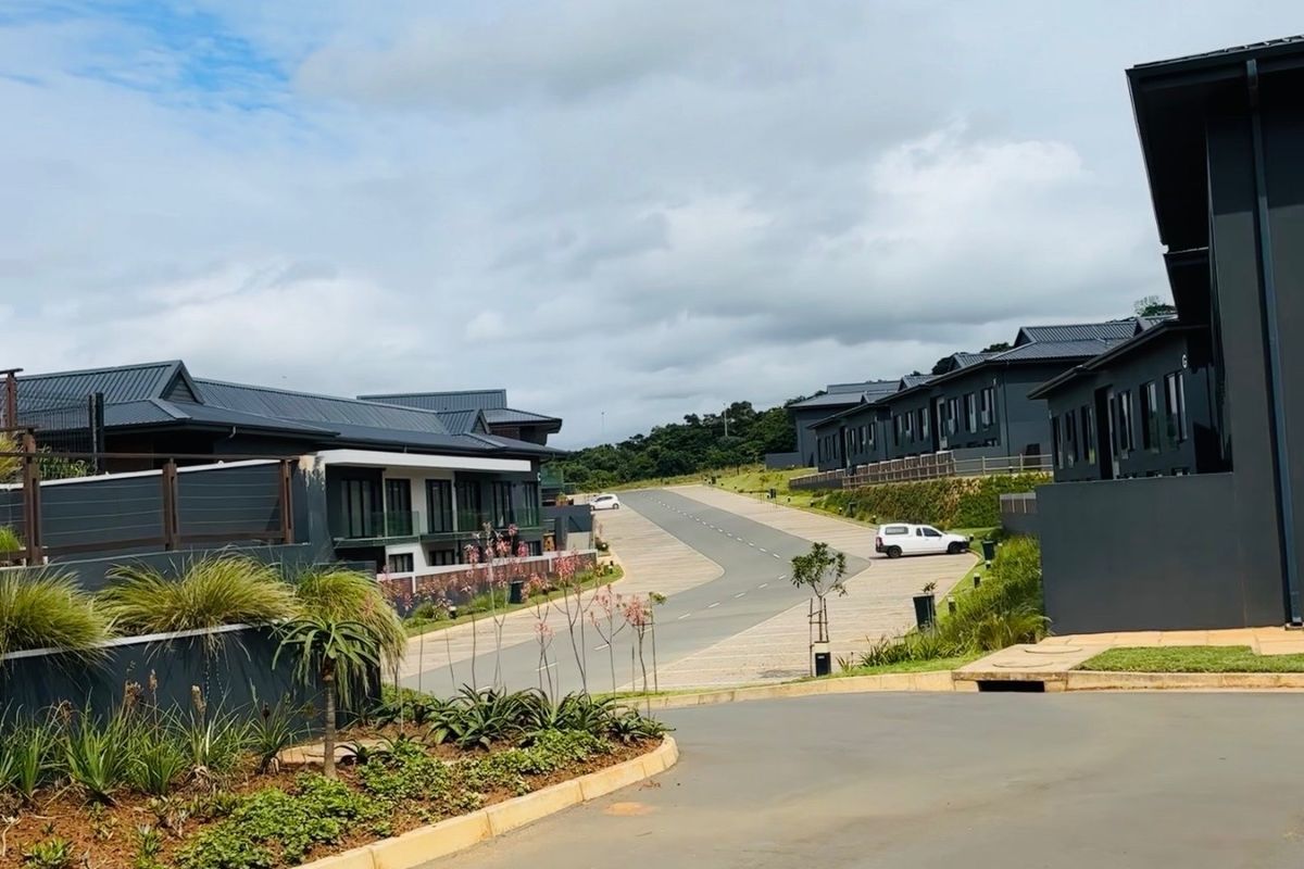 A view of a modern residential complex featuring paved roads, landscaped garden beds, and contemporary apartment buildings with dark metal roofing.