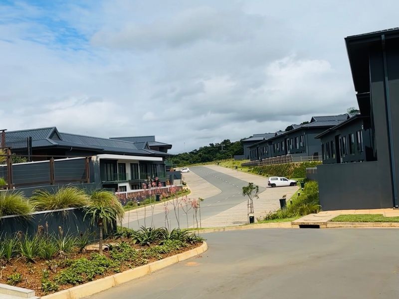 A view of a modern residential complex featuring paved roads, landscaped garden beds, and contemporary apartment buildings with dark metal roofing.