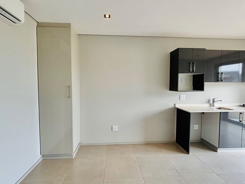 Modern open-plan kitchen featuring sleek dark cabinetry, integrated appliances, tiled flooring, and an air conditioning unit in a clean, minimalist space.