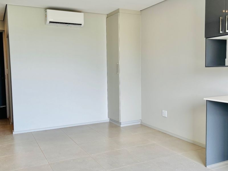A bright, modern open-plan living area featuring neutral tiled flooring, built-in cabinetry, and a wall-mounted air conditioning unit.