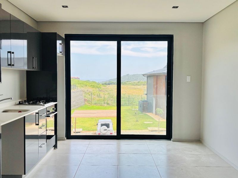 Modern open-plan kitchen featuring sleek dark cabinetry, stone countertops, and integrated appliances, leading out to a garden view through large sliding glass doors.