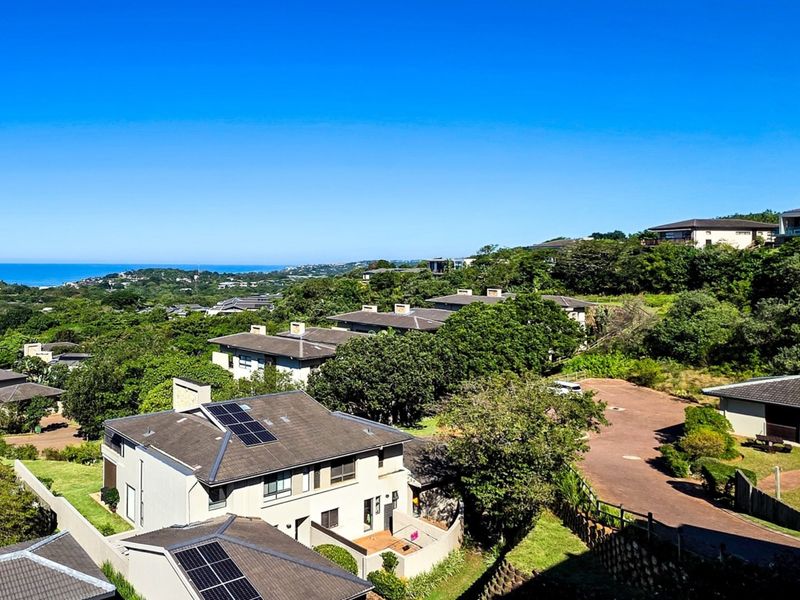 An elevated aerial view of a residential apartment complex featuring tiled roofs, solar panels, and surrounding lush greenery with a distant ocean view.