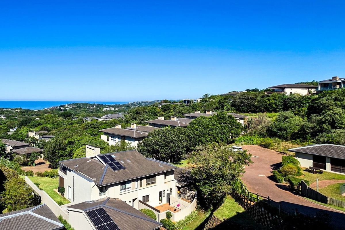 An elevated aerial view of a residential apartment complex featuring tiled roofs, solar panels, and surrounding lush greenery with a distant ocean view.