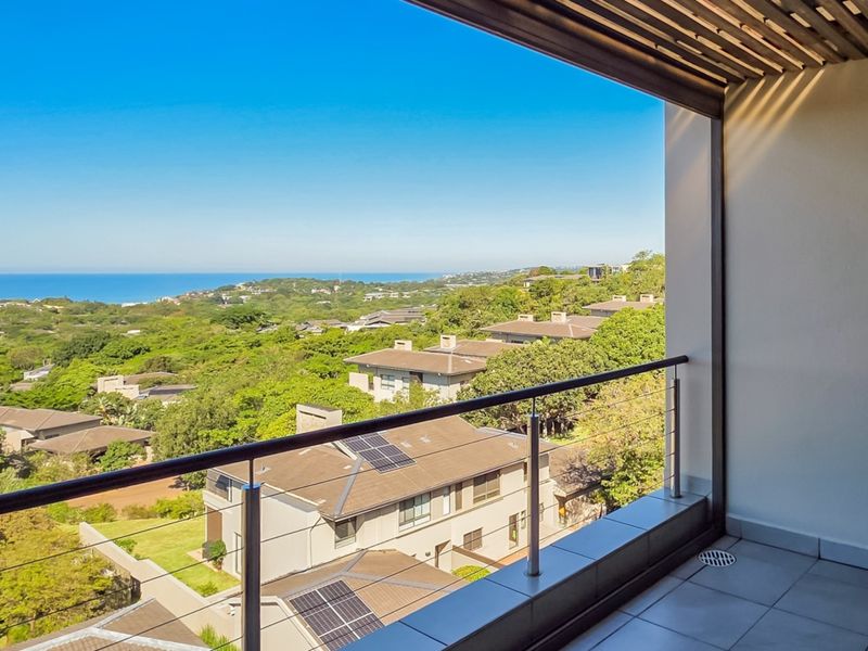 A spacious covered balcony featuring tiled flooring, a modern stainless steel railing, and expansive elevated views of the surrounding landscape and ocean.