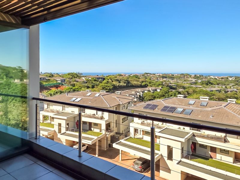 A spacious balcony featuring tiled flooring, a modern stainless steel balustrade, and glass sliding doors, offering an elevated view of the surrounding residential area.