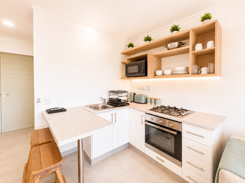 A modern, compact kitchen featuring white cabinetry, light wood open shelving, integrated gas hob, built-in oven, and a breakfast bar extension.