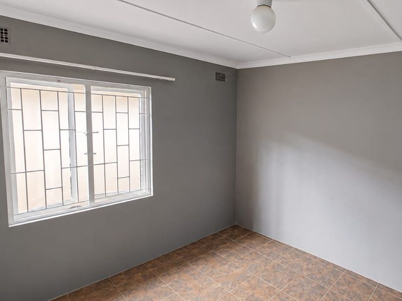 A clean, empty bedroom featuring neutral grey walls, tiled flooring, and a window fitted with security bars.