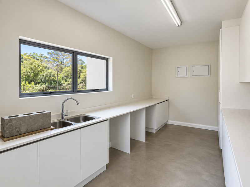 A clean, modern laundry room featuring ample built-in cabinetry, stone countertops, a stainless steel double sink, and large windows overlooking greenery.