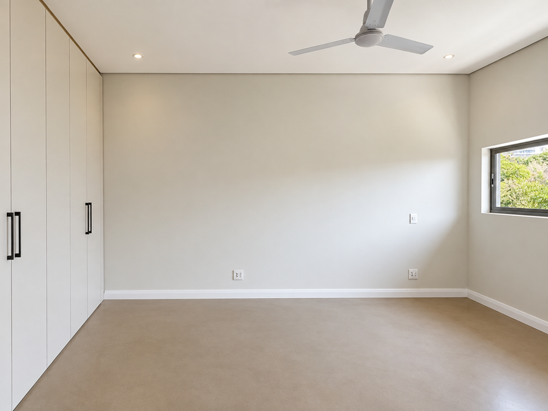 A bright, empty bedroom featuring neutral walls, polished concrete flooring, built-in floor-to-ceiling cupboards, and a modern ceiling fan.