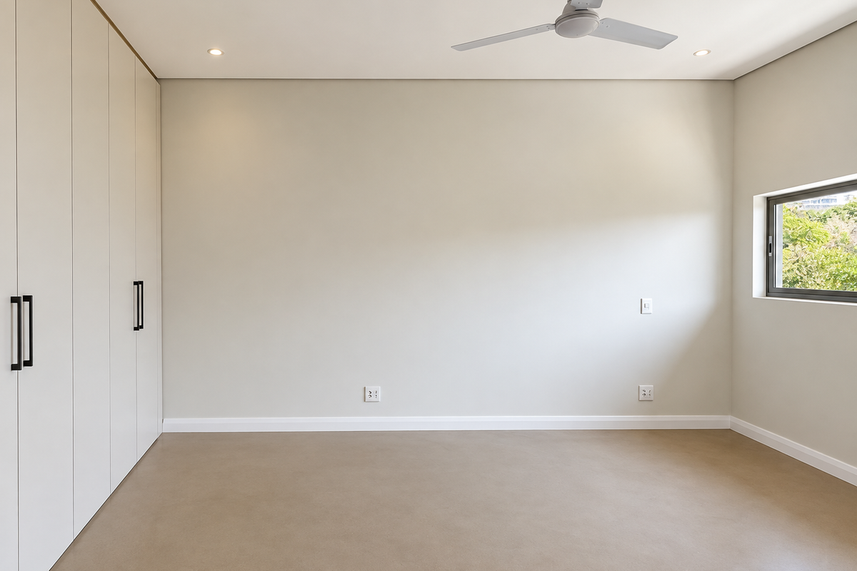 A bright, empty bedroom featuring neutral walls, polished concrete flooring, built-in floor-to-ceiling cupboards, and a modern ceiling fan.