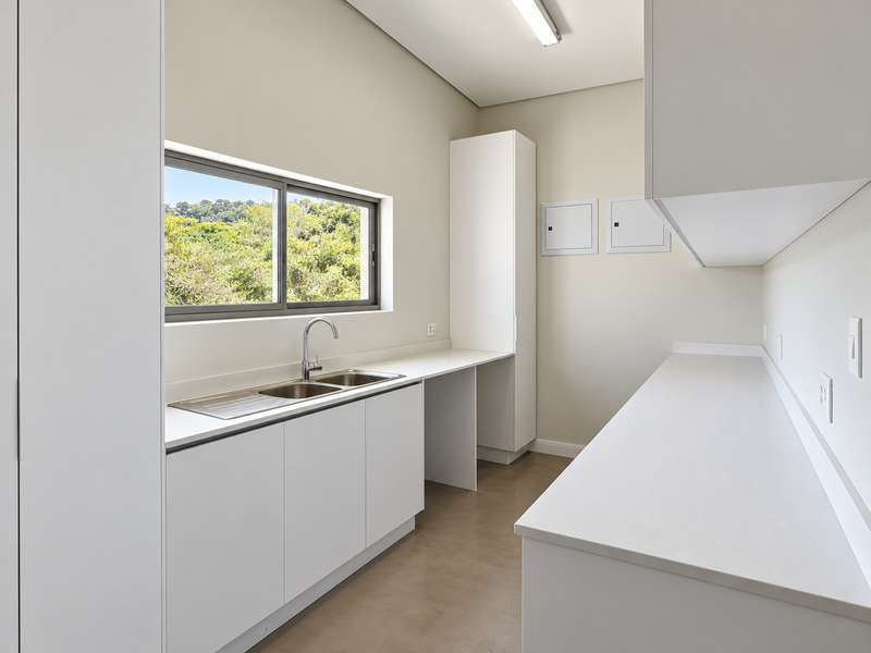 A clean, modern scullery featuring white built-in cabinetry, stone countertops, a double sink, and a window with a pleasant green outlook.