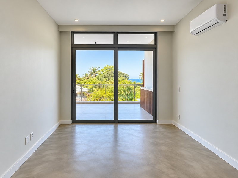 A bright, empty bedroom featuring polished concrete floors, a split-system air conditioner, and large sliding glass doors opening onto a balcony with scenic views.