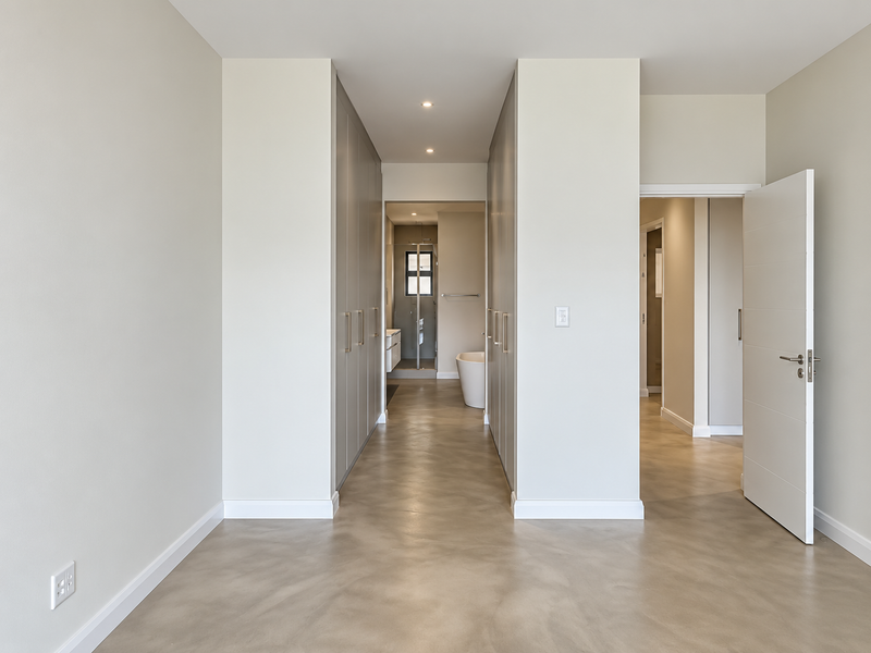 A spacious, modern bedroom featuring polished concrete floors, extensive built-in cabinetry, and a direct view into an en-suite bathroom.