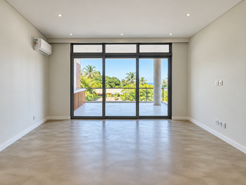 A bright, empty living room featuring polished concrete floors, recessed lighting, a split-system air conditioner, and large sliding doors opening to a balcony.