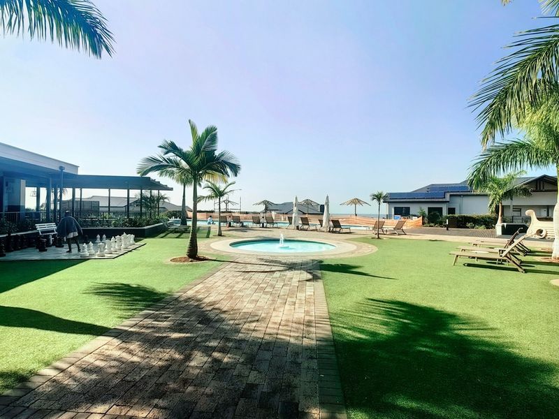 A well-maintained outdoor pool area featuring a central water fountain, paved pathways, artificial lawn, and surrounding palm trees in a residential complex.