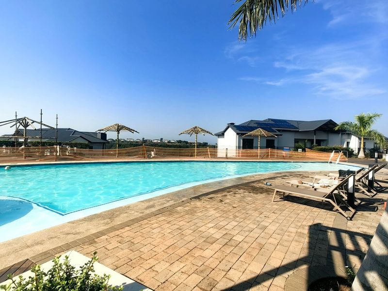Outdoor swimming pool area featuring a paved deck, thatched umbrellas, and temporary safety fencing, set against a clear blue sky.