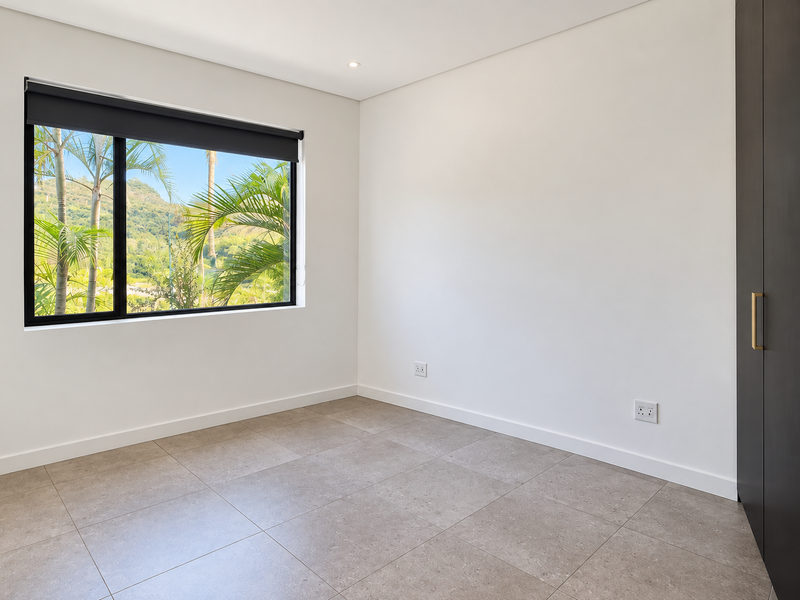 A bright, modern, empty bedroom featuring neutral tiled flooring, built-in dark cabinetry, and a large window with views of the surrounding greenery.