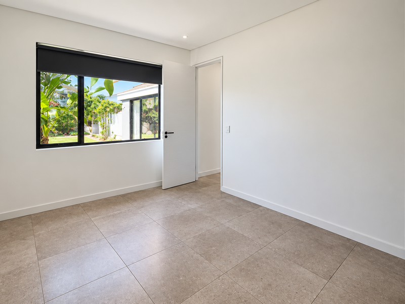A bright, empty bedroom featuring neutral tiled flooring, a built-in wardrobe, and a large window overlooking the garden.
