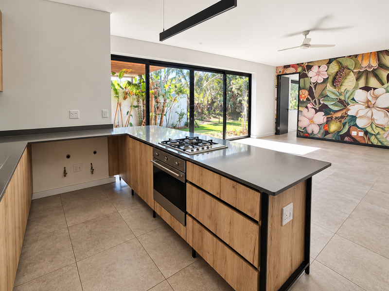 Modern open-plan kitchen and living area featuring wood-look cabinetry, stone countertops, integrated appliances, and large sliding doors opening to a private garden.