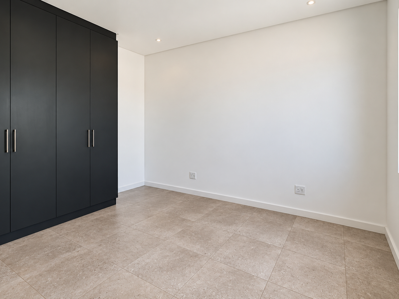 A bright, modern bedroom featuring neutral tiled flooring, clean white walls, and sleek dark built-in cupboards with recessed lighting.
