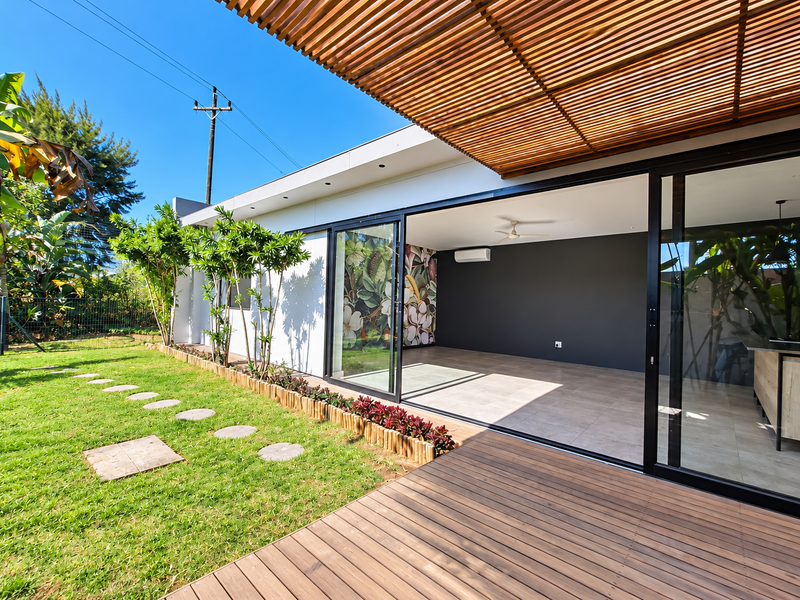 A modern covered timber deck featuring a slatted pergola, seamless indoor-outdoor flow through large sliding glass doors, and a well-maintained garden.