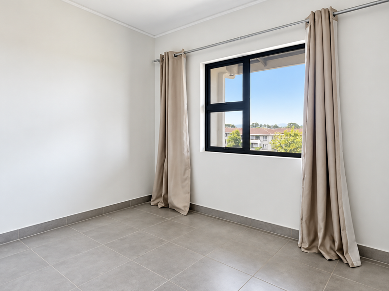 A bright, empty bedroom featuring neutral tiled flooring, clean white walls, and a large window with a view of the surrounding neighborhood.