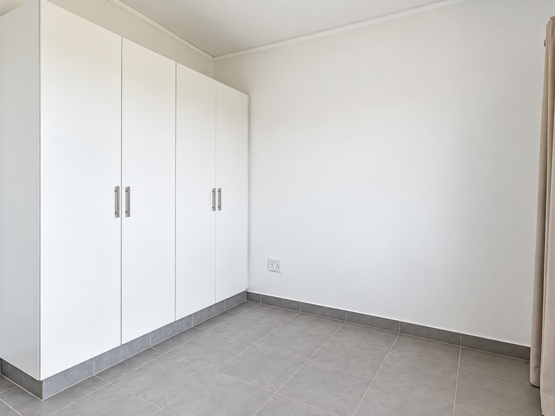 A bright, empty bedroom featuring neutral tiled flooring, clean white walls, and modern built-in cupboards with sleek hardware.
