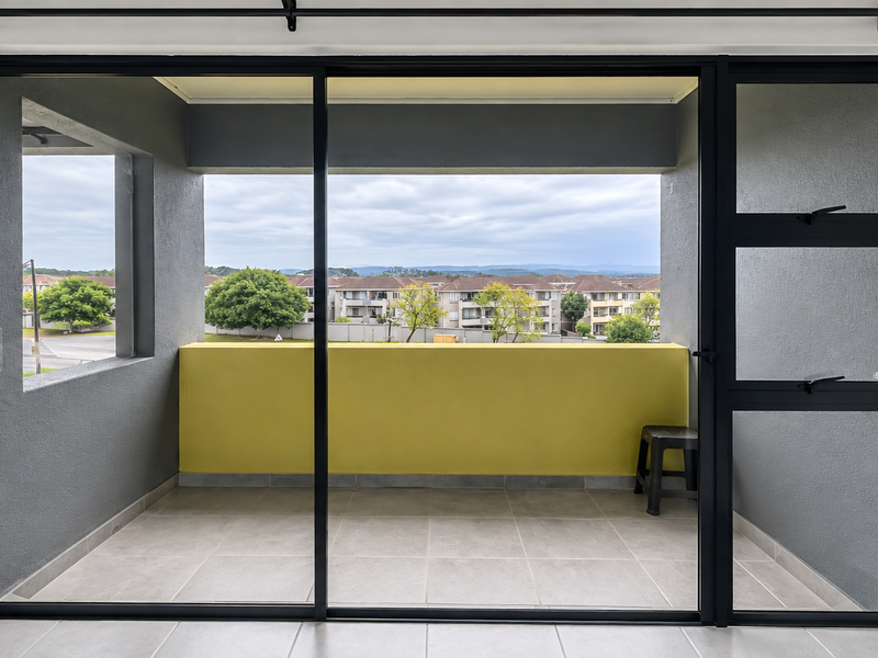 A clean, modern balcony featuring tiled flooring, dark-framed sliding glass doors, and a vibrant yellow accent wall with views of the surrounding neighborhood.