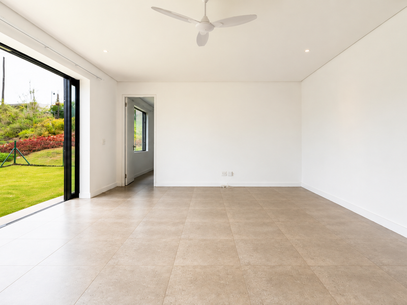 A bright, empty living room featuring large sliding glass doors that open to a garden, tiled floors, a ceiling fan, and air conditioning.