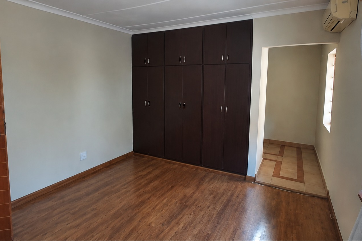 A clean, empty bedroom featuring dark wood-finish built-in cupboards, laminate flooring, and an integrated air conditioning unit.