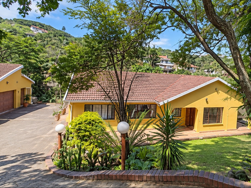 Front view of a yellow house featuring a tiled roof, paved driveway, double garage, and a landscaped garden with a brick retaining wall.