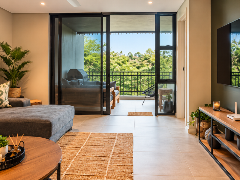 A modern, well-lit living room featuring large sliding glass doors that open onto a private balcony with lush green views.