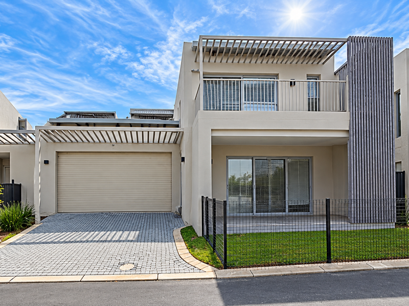 Modern townhouse exterior featuring a paved driveway, automated garage, balcony with pergola, and a private front garden area with secure metal fencing.